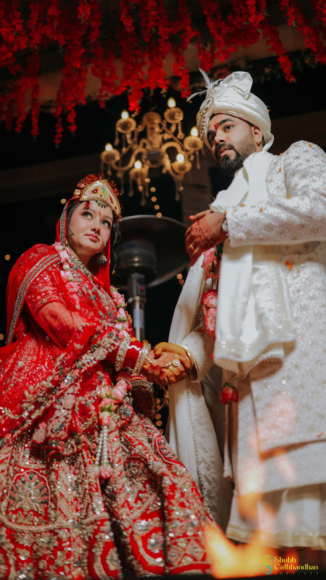 The couple holding hands beneath a canopy of red flowers and chandeliers
